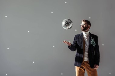 handsome man in glasses throwing up disco ball on grey background clipart