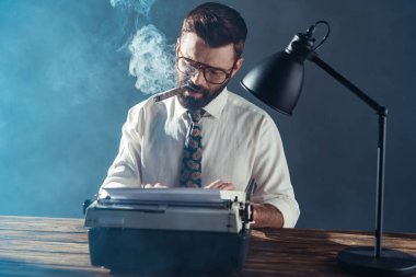 handsome journalist in glasses sitting at table, typing on vintage typewriter and smoking on grey background clipart