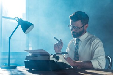 bearded journalist in glasses sitting at table, holding cigar and looking at vintage typewriter near window clipart