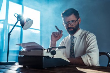 bearded journalist in glasses sitting at table with vintage typewriter, holding cigar and looking at camera near window clipart