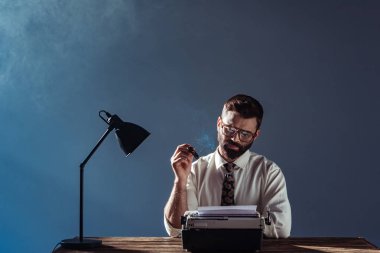 bearded journalist sitting at table with lamp, looking at vintage typewriter and smoking on grey background clipart