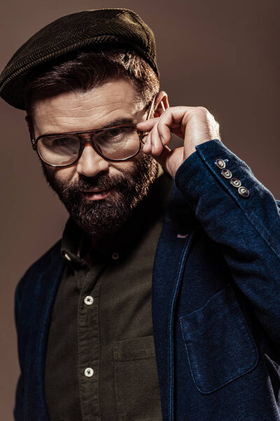 handsome bearded man touching glasses and looking at camera isolated on brown