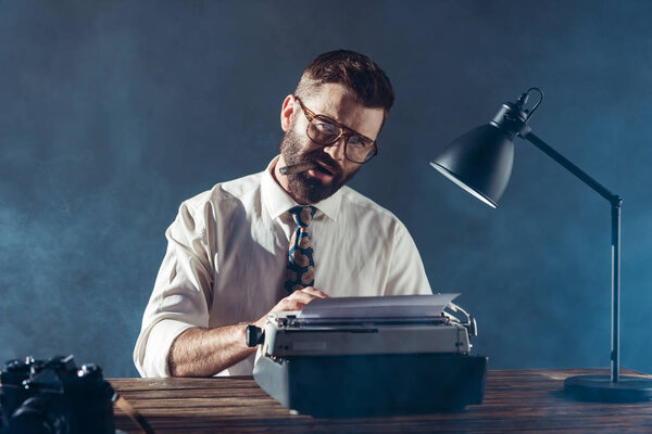 bearded journalist sitting at table, typing on typewriter, smoking and looking at camera on grey background