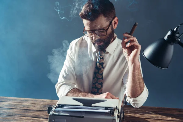 穿着眼镜坐在桌旁的英俊记者 看着老式打字机 在灰色的背景上抽烟 — 图库照片