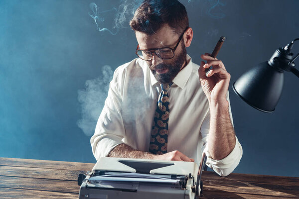 handsome journalist in glasses sitting at table, looking on vintage typewriter and smoking on grey background