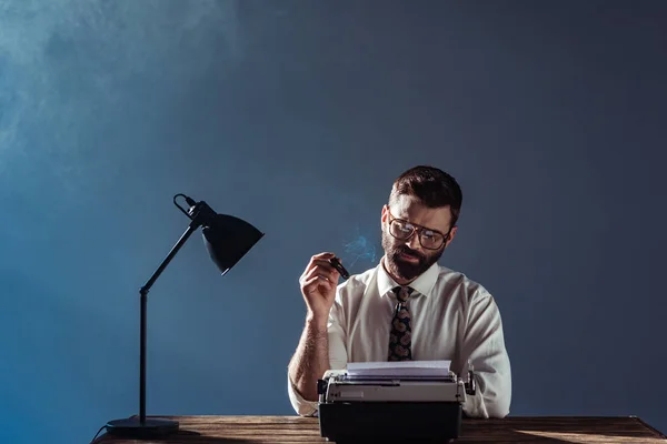 胡须记者坐在桌旁与灯 看着老式打字机和吸烟在灰色的背景 — 图库照片