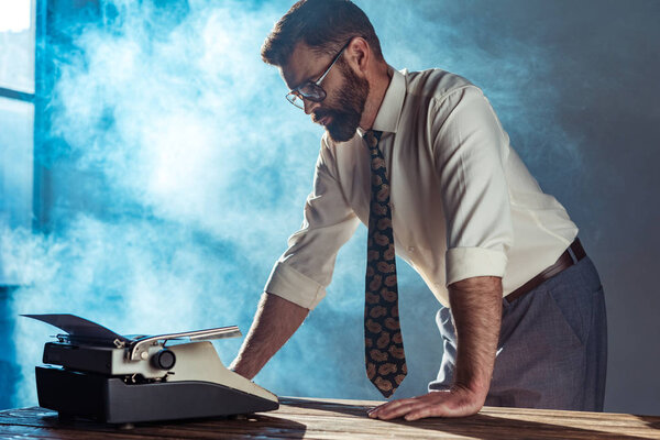 handsome bearded man holding cigar and looking at typewriter