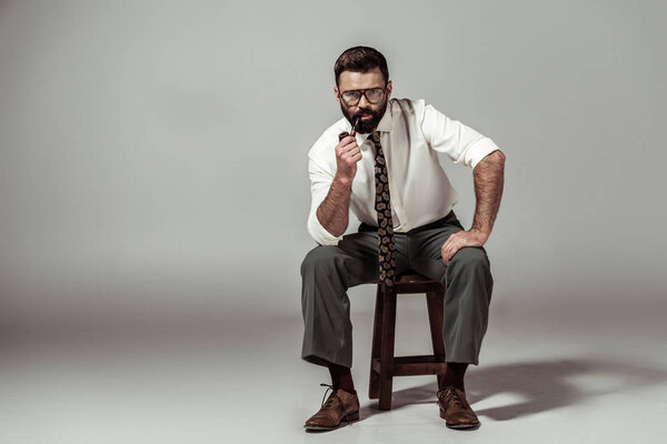 handsome bearded man in glasses sitting on chair and looking at camera while smoking pipe
