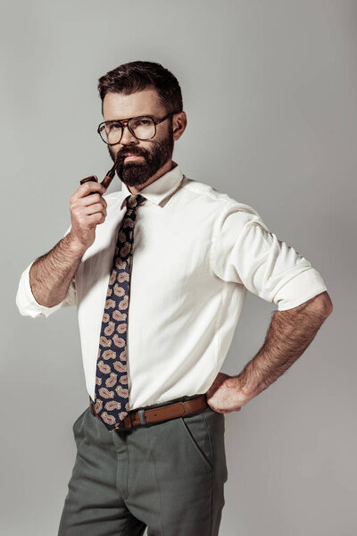 handsome bearded man in glasses and shirt smoking pipe and looking at camera isolated on grey