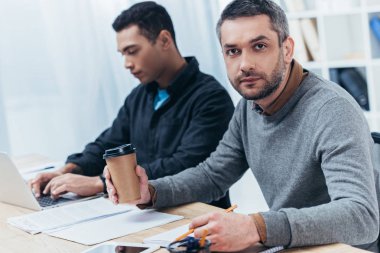 Kağıt bardak tutan ve genç erkek meslektaşı Office ile çalışırken kameraya bakarak ciddi iş adamı