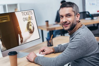 Bilet online web sitesi ile masaüstü bilgisayar çalışma ve bu öğeleri office kamerada gülümseyen yakışıklı sakallı işadamı