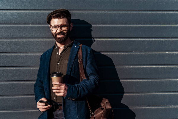 handsome bearded man in glasses and cap holding paper cup and smiling at camera