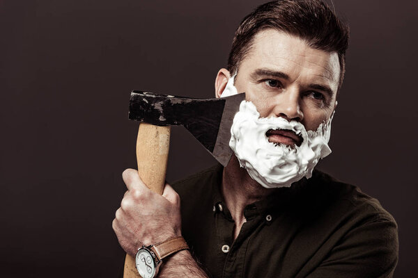 handsome bearded man in brown shirt shaving with ax isolated on brown