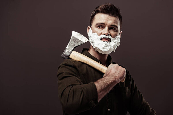 handsome bearded man with shaving foam on face holding ax isolated on brown