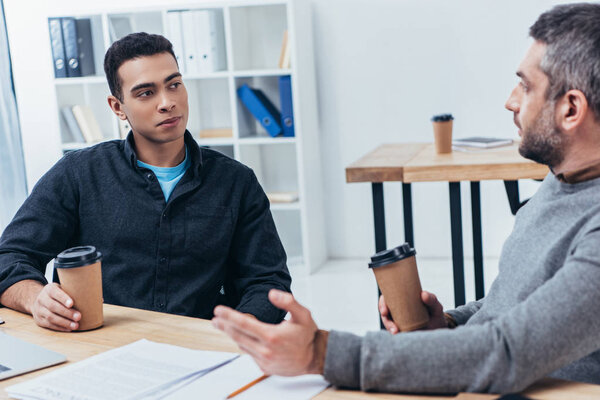 professional businessmen holding paper cups and talking while sitting at workplace