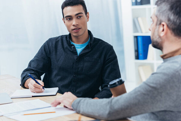 young businessman writing in notebook and looking at colleague sitting in office