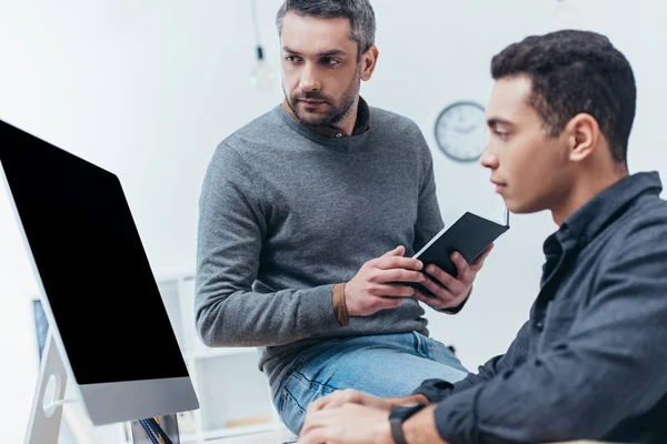 serious business mentor holding notebook and looking at young colleague using desktop computer at workplace