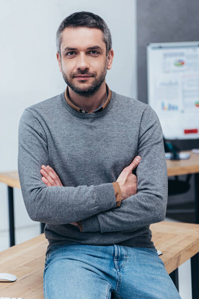 handsome bearded businessman sitting on table with crossed arms and looking at camera