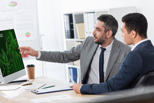 bearded businessman showing desktop computer with trading graphs on screen to young colleague at workplace