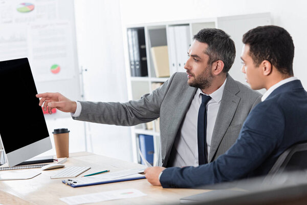 bearded businessman showing desktop computer with blank screen to young colleague at workplace