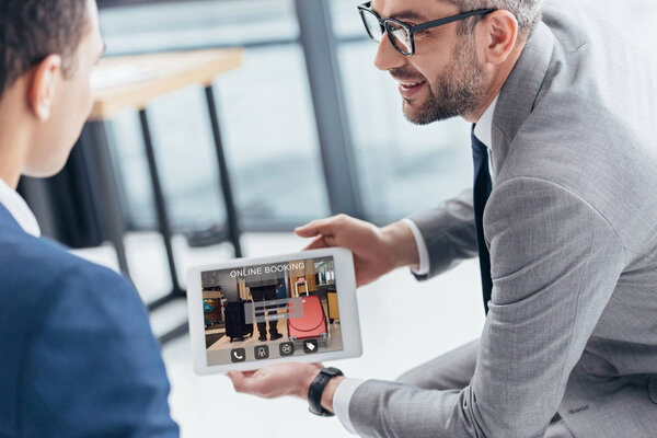 cropped shot of businessman in eyeglasses showing digital tablet with online booking app on screen to male colleague in office