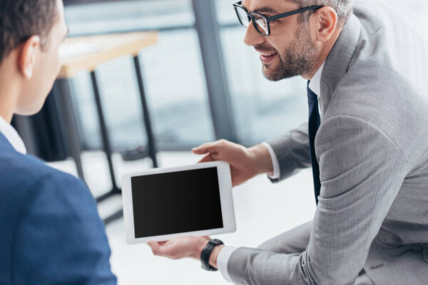 cropped shot of businessman in eyeglasses showing digital tablet with blank screen to male colleague in office