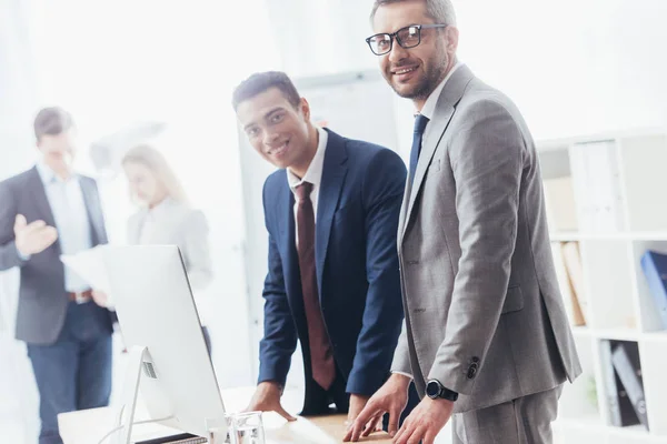 Hombres Negocios Felices Sonriendo Cámara Mientras Apoyan Mesa Con Computadora — Foto de Stock