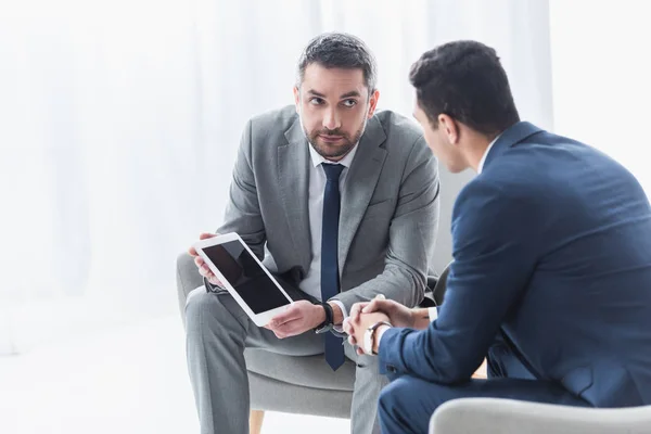 serious business mentor holding digital tablet with blank screen and looking at young colleague in office