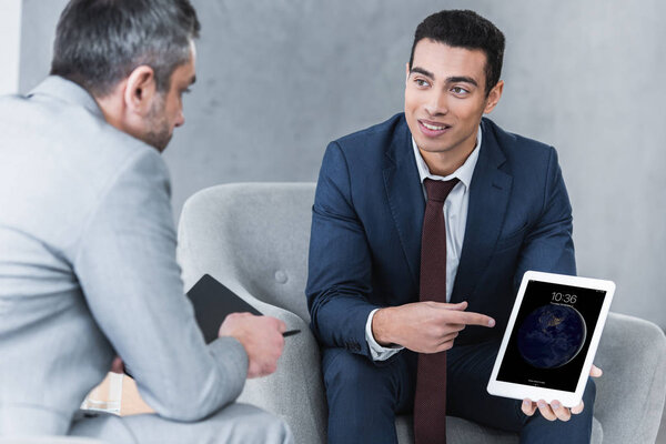 smiling young businessman pointing at digital tablet and looking at male colleague during conversation