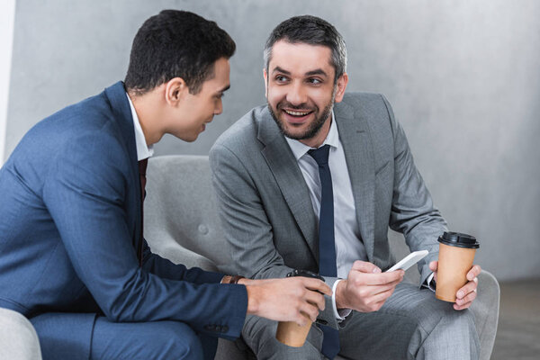 smiling businessmen drinking coffee from paper cups and using smartphone together 