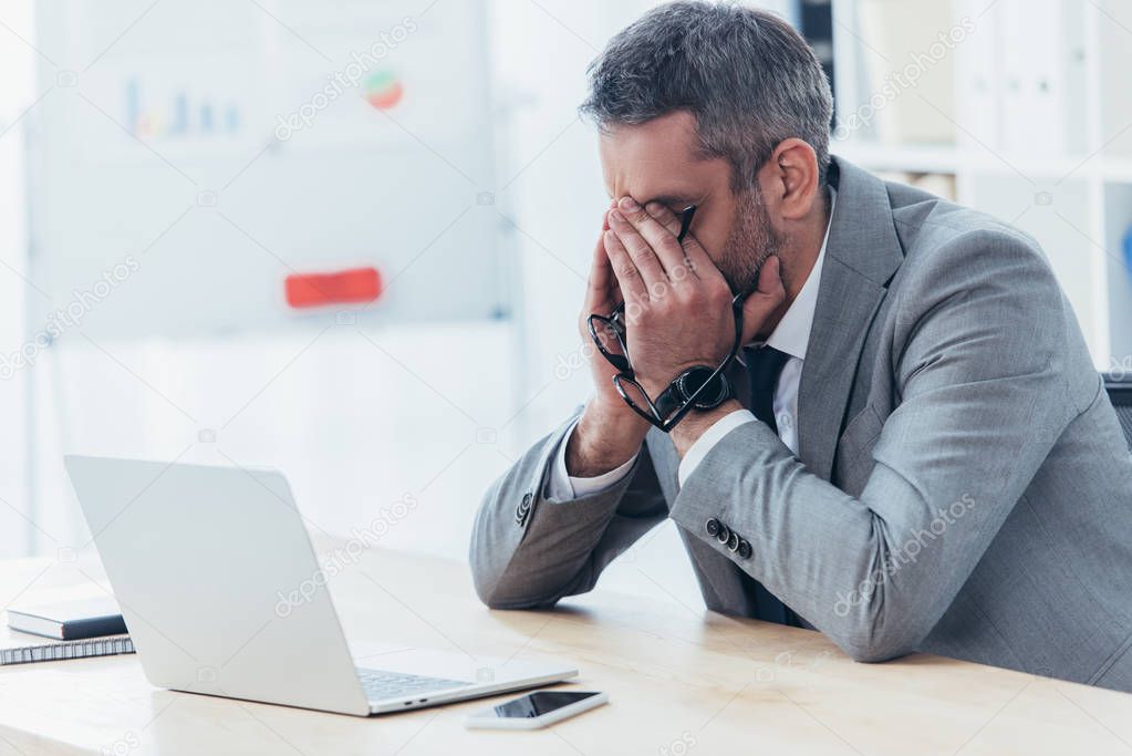 tired businessman holding eyeglasses and rubbing eyes while sitting at workplace