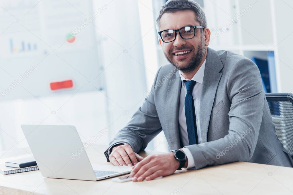 cheerful businessman in eyeglasses using laptop and smiling at camera in office