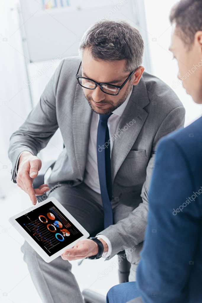 cropped shot of businessman in eyeglasses showing digital tablet with business charts to male colleague in office