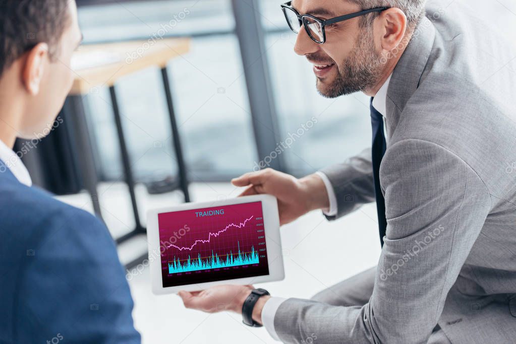 cropped shot of businessman in eyeglasses showing digital tablet with trading graphs on screen to male colleague in office