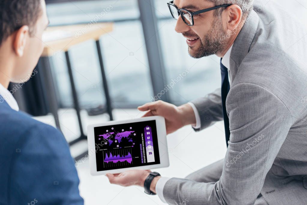 cropped shot of businessman in eyeglasses showing digital tablet with charts and graphs on screen to male colleague in office