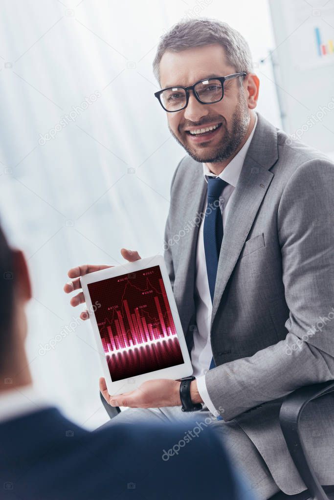 smiling businessman in eyeglasses holding digital tablet with business charts on screen