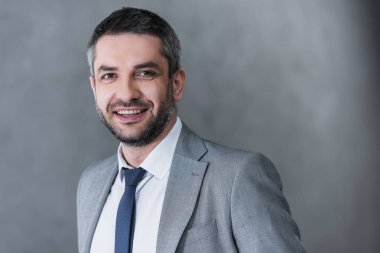 portrait of handsome businessman in formal wear smiling at camera on grey  clipart