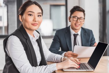 kağıtları ve dizüstü Office ile çalışırken kameraya gülümseyen genç Asya iş adamları