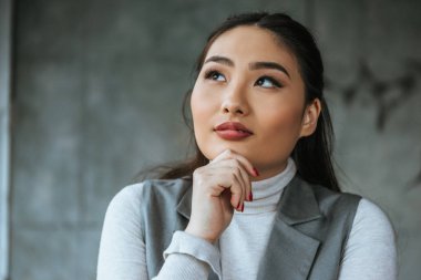 pensive young asian woman holding hand on chin and looking up in office clipart