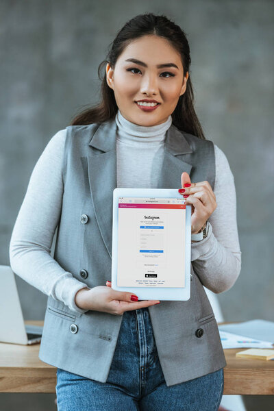 young asian businesswoman holding digital tablet with instagram application and smiling at camera