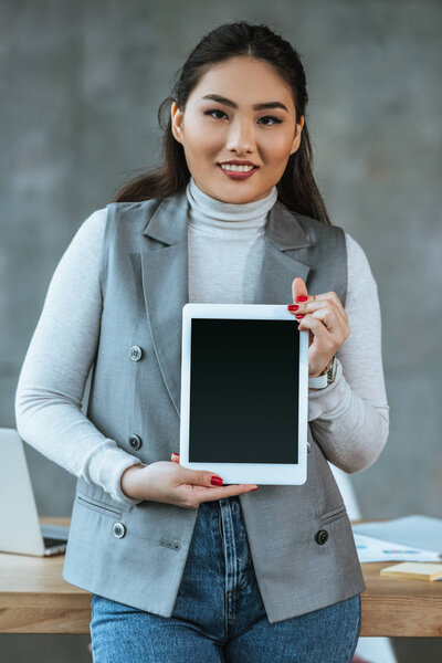young asian businesswoman holding digital tablet with blank screen and smiling at camera in office