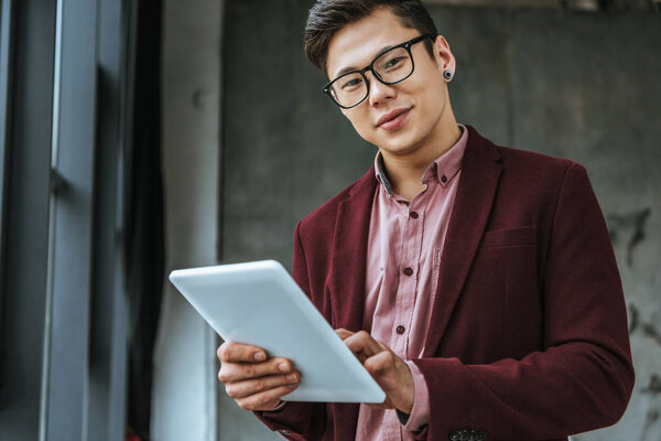 handsome kazakh businessman in eyeglasses using digital tablet and looking at camera in office 