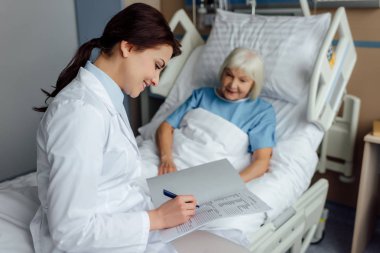 Yatağın üstüne oturan ve tanı hastane yatağında yatan kadın kıdemli süre yazma gülümseyen kadın doktor