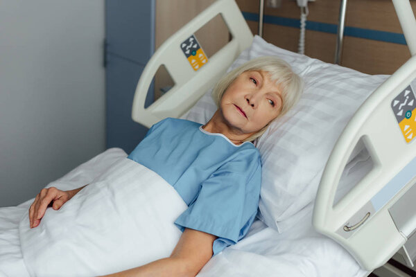 sad senior woman lying in bed in hospital and looking away