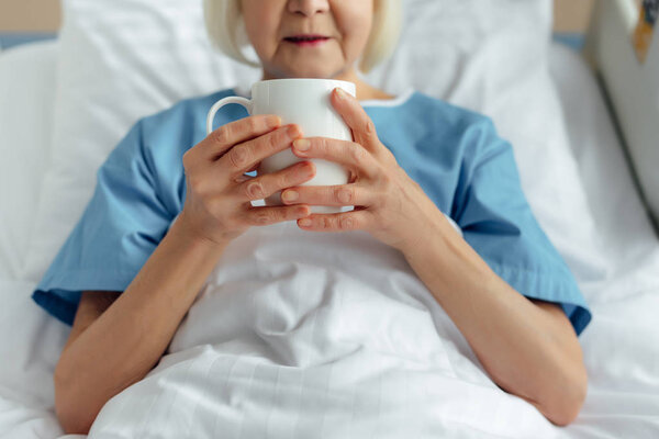 cropped view of senior woman lying in bed and drinking tea in hospital