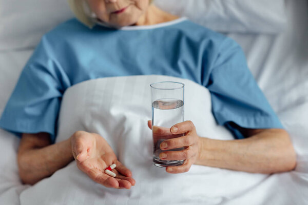 partial view of senior woman lying in bed and holding pills with glass of water in hospital