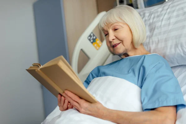 微笑的灰色头发的老年妇女躺在床上看书在医院 — 图库照片