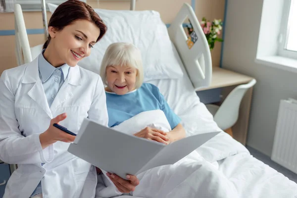 美丽的微笑的女医生拿着诊断和咨询老年妇女躺在病床上 — 图库照片