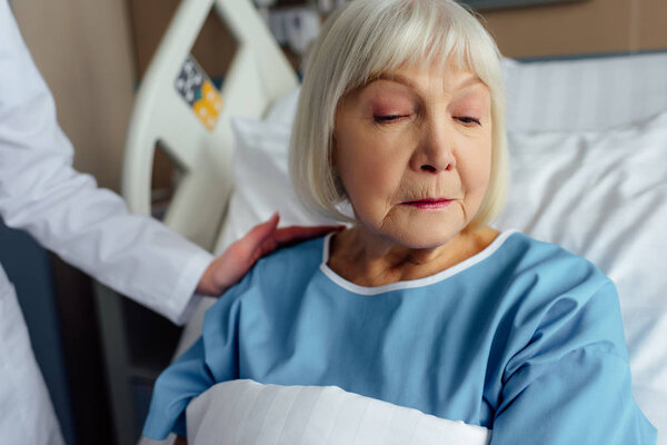female doctor consoling upset senior woman with grey hair lying in hospital bed