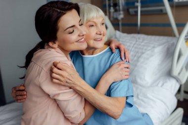 smiling senior woman and daughter sitting on bed and embracing in hospital clipart